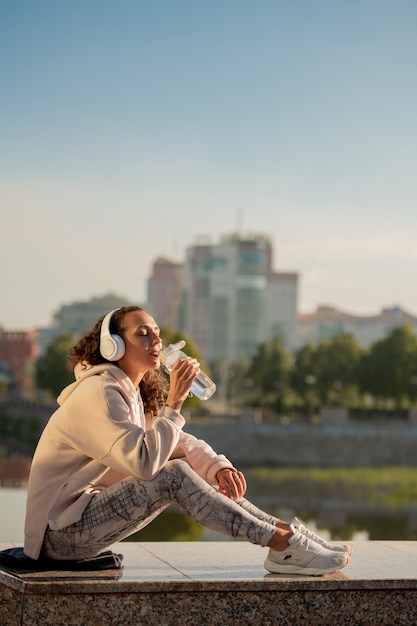 Foto mujer sedienta descansada en ropa deportiva bebiendo agua de la botella mientras está sentada contra la orilla del río después de entrenar al aire libre y disfrutar de la música