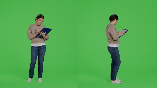 Mujer segura mirando archivos de portapapeles en pantalla verde de cuerpo completo, tomando notas en papeles y trabajando en análisis. Modelo femenino escribiendo información sobre documentos en estudio.