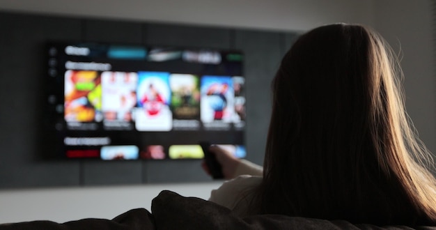 Mujer selecciona canales de televisión con control remoto en la sala de estar en el sofá Mujer controla la televisión usando un control remoto moderno