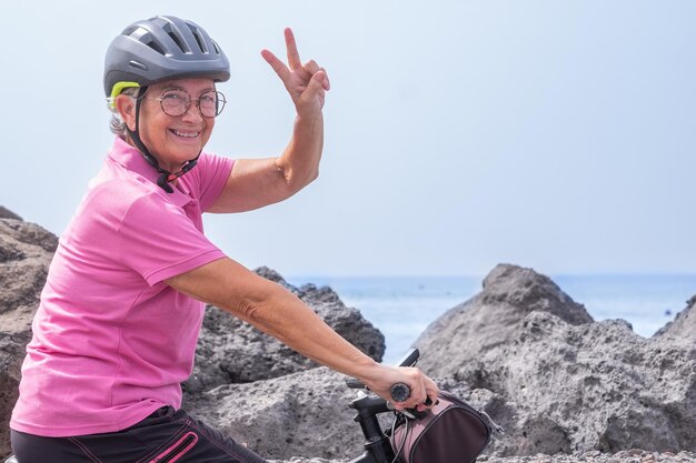 Mujer senior feliz activa con casco corriendo en su bicicleta eléctrica a lo largo del mar mirando a la cámara con el signo de la victoria Concepto de libertad y vacaciones