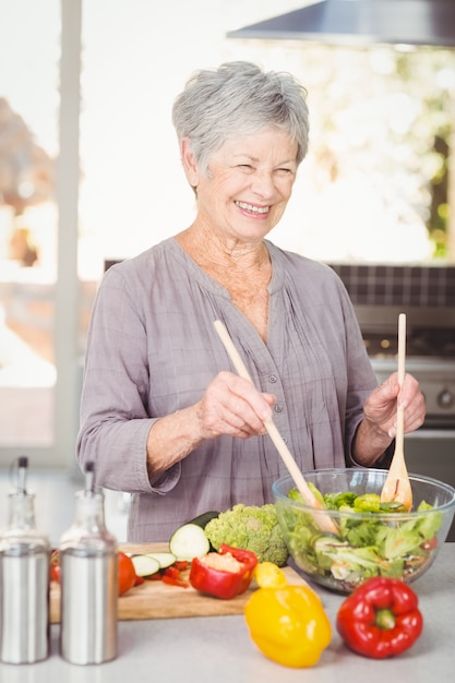 Mujer senior feliz lanzando ensalada mientras está de pie en la cocina