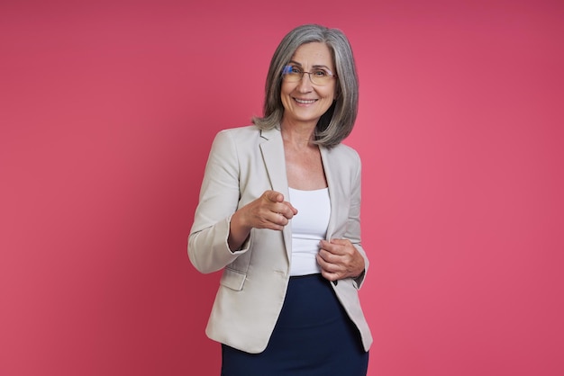 Foto mujer senior feliz en ropa formal apuntando a la cámara mientras está de pie contra un fondo rosa