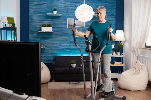Mujer senior no válida haciendo aeróbicos en bicicleta en la sala de estar para adelgazar. Pensionista de jubilación viendo videos cardiovasculares en línea en la televisión haciendo ejercicio de los músculos de las piernas