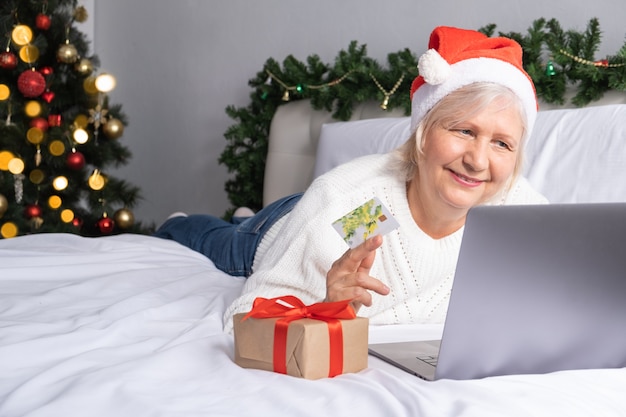 Mujer senior positiva haciendo compras para Navidad en casa en la cama.