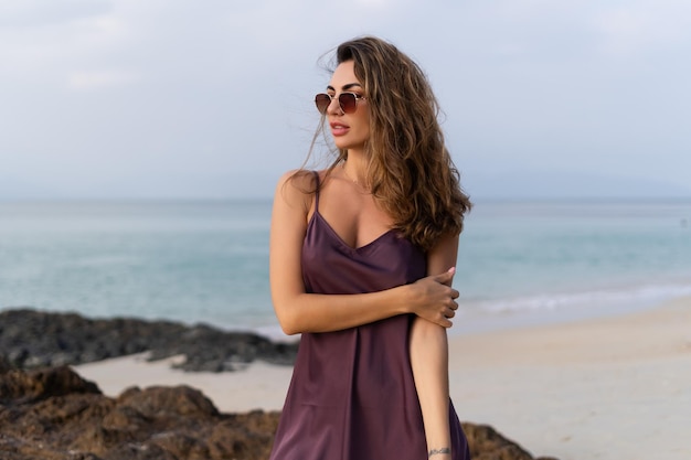 Mujer sensual tierna romántica con estilo en vestido de seda y gafas de sol en la playa al atardecer