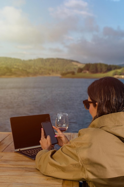 Mujer sentada afuera frente a su computadora portátil mientras revisa su teléfono móvil con lago y montañas en el fondo