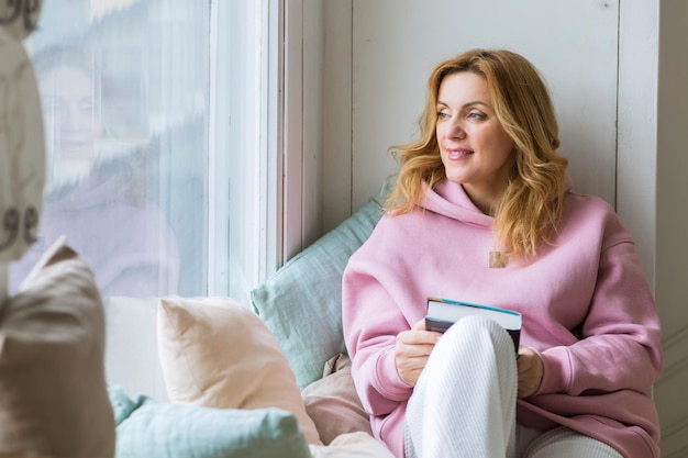 Mujer sentada en el alféizar de la ventana y leyendo un libro. Desintoxicación de las redes sociales. Estilo de vida lento. Mujer feliz disfrutando de la literatura. Tranquilo ratón de biblioteca pasando el día libre de gadgets