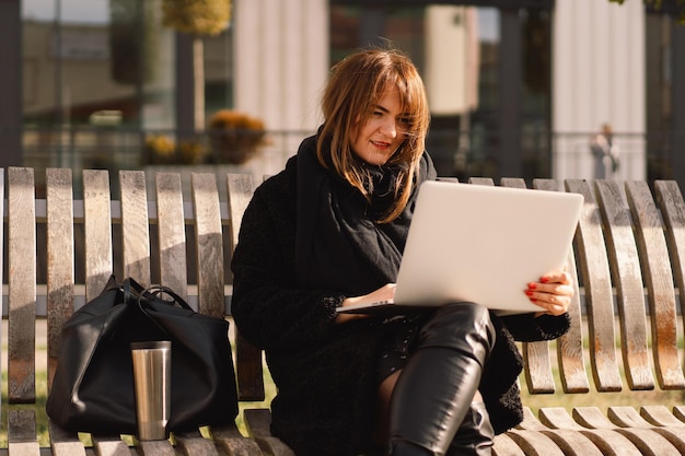 Una mujer sentada en un banco en la ciudad, usa una computadora. Mujer autónoma. Personas y tecnología