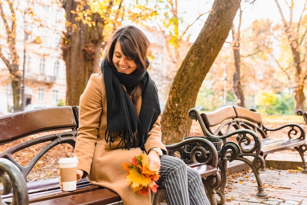 Mujer sentada en el banco en el parque de la ciudad de otoño tomando café