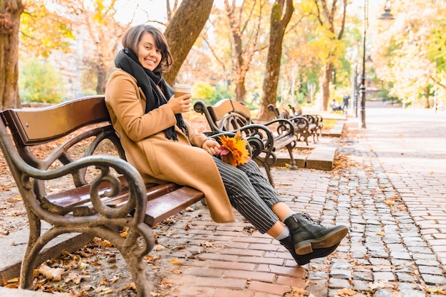 Mujer sentada en el banco en el parque de la ciudad de otoño tomando café