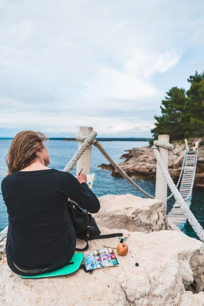 Mujer sentada en el borde dibujando un paisaje marino