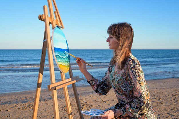 Foto mujer sentada en un caballete pintando en la orilla del mar