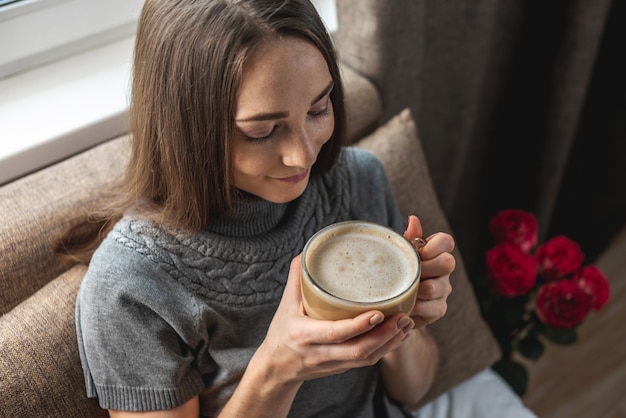 Mujer sentada en la cama junto a la ventana y disfrutando de un café en un ambiente acogedor