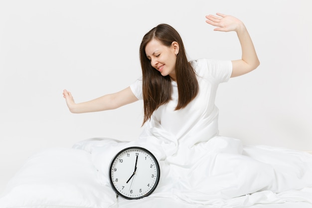 Mujer sentada en la cama con reloj redondo, sábana blanca, almohada, envolviendo en una manta en la pared blanca