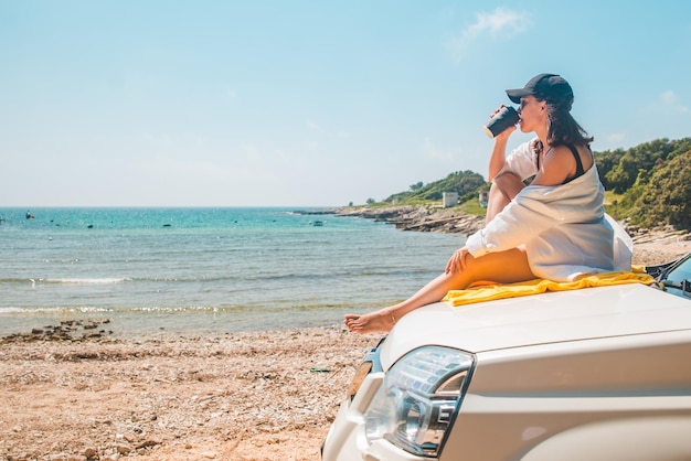 Mujer sentada en el capó del coche bebiendo café disfrutando de la vista del mar