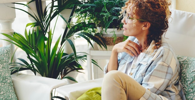 Una mujer sentada en casa en el jardín con plantas verdes alrededor y mirando. Concepto de estilo de vida único de personas femeninas pensativas. Señora adulta pensando y disfrutando relajarse. Hermosa mujer hipster