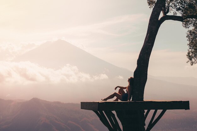 Mujer sentada cerca de un árbol contra el telón de fondo de una silueta de un volcán