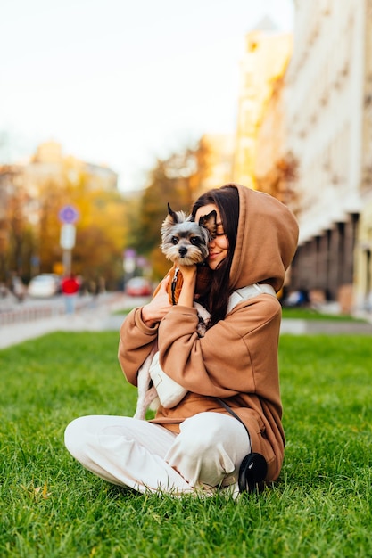 mujer sentada en el césped con un lindo perrito en sus brazos en un fondo de la calle y jugando