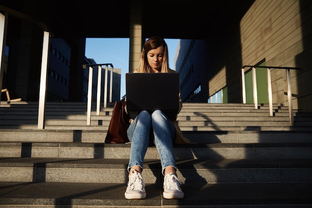 Mujer sentada con una computadora portátil en pasos al aire libre contra la puesta de sol