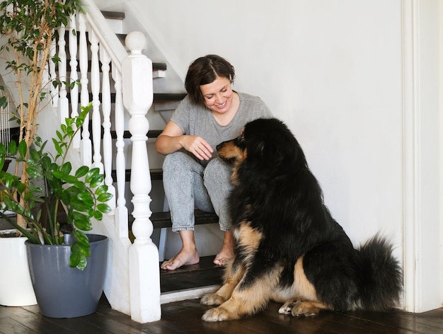 Mujer sentada en las escaleras en casa, usando un teléfono inteligente con un perro grande negro