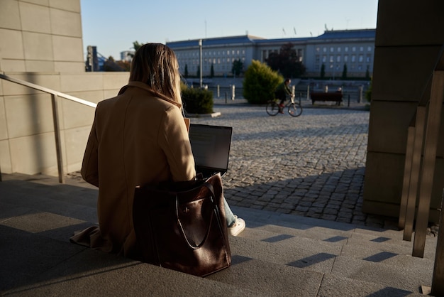 Mujer sentada en escalones al aire libre y usando el concepto de computadora portátil de trabajo remoto y educación en línea