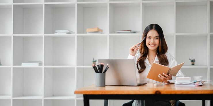 Una mujer sentada en un escritorio con una computadora portátil y un cuaderno Ella está sonriendo y sosteniendo un bolígrafo La escena sugiere un ambiente de trabajo productivo y positivo
