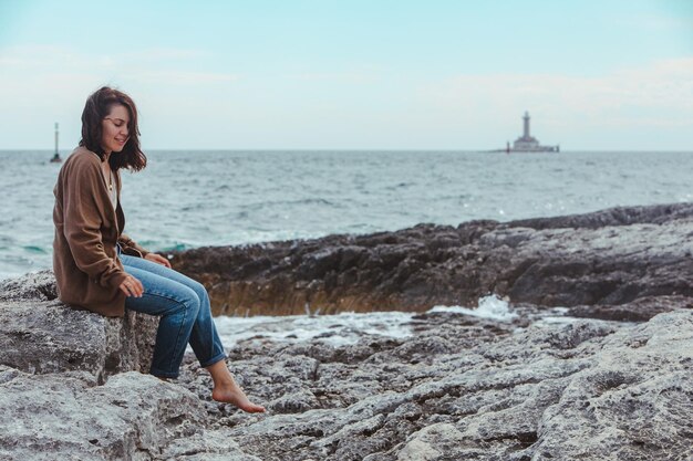 Mujer sentada junto a la playa de mar rocoso en el faro de jeans mojados en el fondo