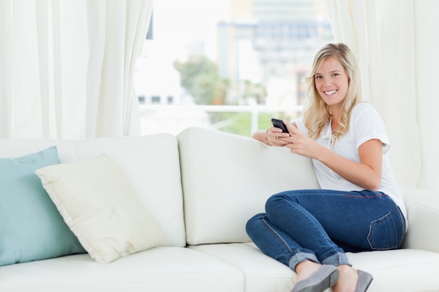 Una mujer sentada de lado en el sofá mientras usa su teléfono y mira a la cámara