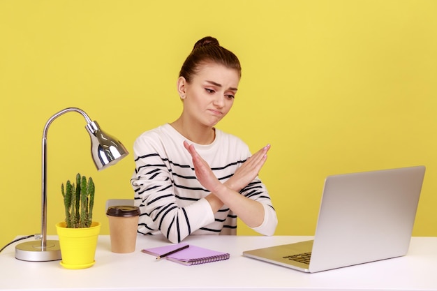 Mujer sentada en el lugar de trabajo cruzando las manos mostrando el signo x mirando la pantalla del portátil con el ceño fruncido