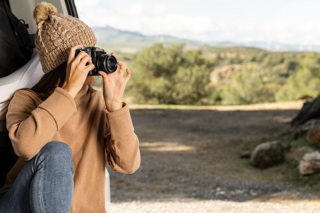 Mujer sentada en el maletero del coche durante un viaje por carretera y sosteniendo la cámara