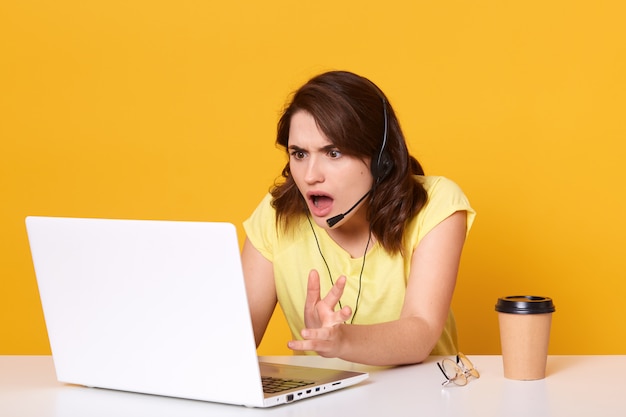 Foto mujer sentada en la mesa y mirando la pantalla de su portátil con consternación y horror, manteniendo la boca abierta, vistiendo una camiseta casual, posando aislado en amarillo, trabajando en línea.
