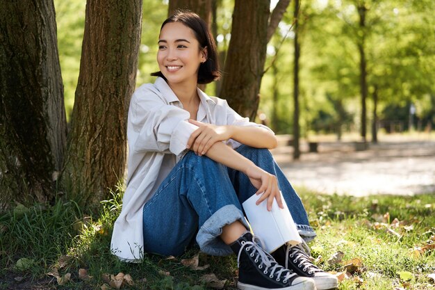 Mujer sentada en el parque con su libro favorito apoyándose en el árbol bajo la sombra en un día soleado disfrutando de natu