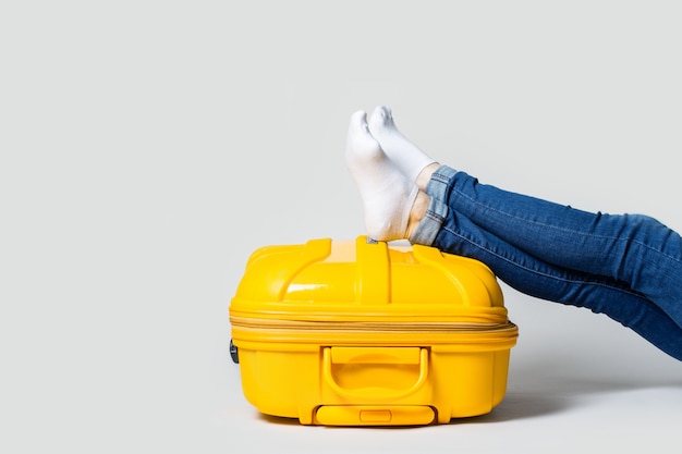 Mujer sentada con piernas en maleta de plástico amarillo en un espacio de luz. Concepto de viaje, expectativa de vuelo, vacaciones.