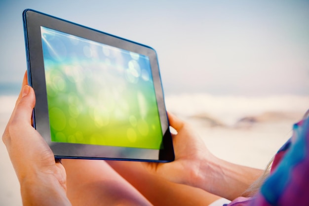 Mujer sentada en la playa en una tumbona usando una tableta que muestra un diseño abstracto verde y azul