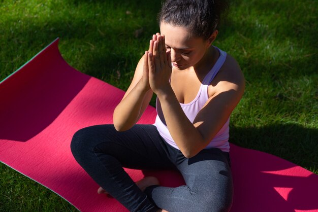 Mujer sentada en las posiciones de loto con los ojos cerrados haciendo yoga en la estera de yoga en la hierba verde en el parque en un día cálido
