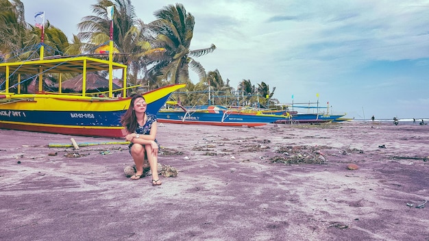 Foto mujer sentada en una roca contra los barcos en la playa
