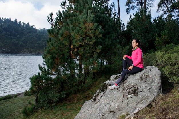 Mujer sentada en una roca a la orilla de un lago con arboles al fondo