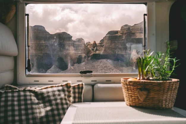 Una mujer sentada en las rocas vista desde el interior de una autocaravana moderna a través de la ventana Concepto de estilo de vida nómada y destino de viaje escénico Casa alternativa y vida fuera de la red
