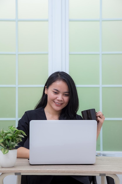 Mujer sentada en una silla con una computadora portátil en la mesa sosteniendo un vaso y mirando la pantalla