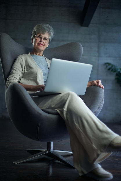 Mujer sentada en una silla moderna cerca de la ventana en una habitación luminosa y acogedora en casa, está trabajando en una laptop en...