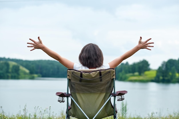 Mujer sentada en una silla en la naturaleza cerca del lago Relajación al aire libre en verano Levantando las manos