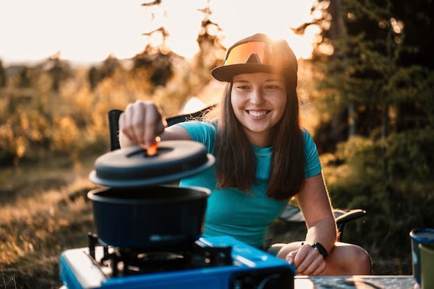 Mujer sentada en sillas fuera de la tienda Acampar al atardecer en el bosque Actividad recreativa al aire libre Cocinar la cena con equipo de campamento en el campamento Viajes de verano Aventura al aire libre