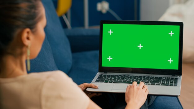 Mujer sentada en el sofá en la acogedora sala de estar trabajando en una computadora portátil con una pantalla verde simulada chroma key. Freelancer que usa una PC aislada, navega en Internet para buscar publicidad en las redes sociales