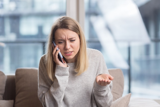 Mujer sentada en el sofá en casa con un fuerte dolor de cabeza tratando de llamar a un médico con un teléfono