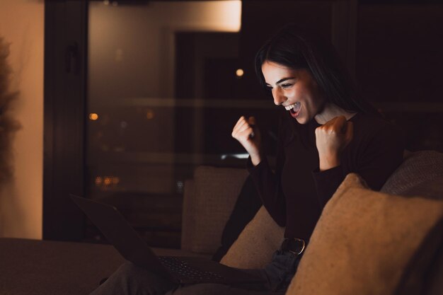 Mujer sentada en el sofá leyendo correo electrónico en la computadora portátil hace que el gesto sí se sienta feliz Emprendedora recibe grandes noticias de negocios celebra el crecimiento profesional avance logro ganar momento de victoria de subasta en la noche xA