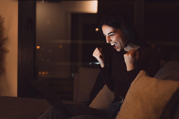 Mujer sentada en el sofá leyendo correo electrónico en la computadora portátil hace que el gesto sí se sienta feliz Emprendedora recibe grandes noticias de negocios celebra el crecimiento profesional avance logro ganar momento de victoria de subasta en la noche xA