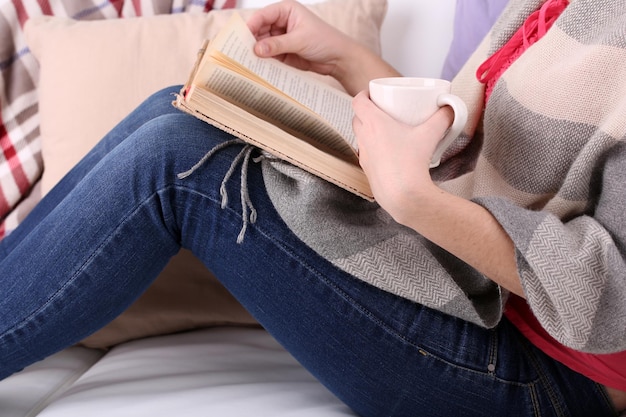 Mujer sentada en el sofá leyendo un libro y bebiendo café o té de cerca