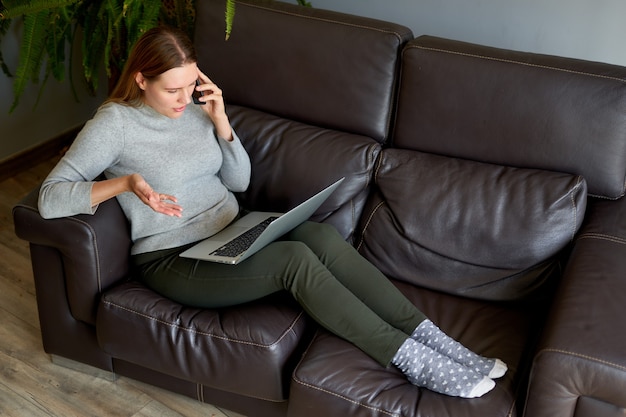 Mujer sentada en el sofá con el portátil y hablando por teléfono en casa. Estudiante universitario que estudia en la computadora portátil y que usa el teléfono.