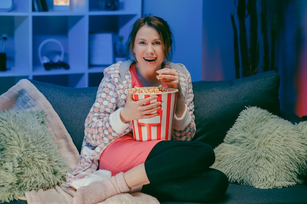 Mujer sentada en el sofá del sofá de la sala viendo un programa interesante divertido y señalando compartir con comer palomitas de maíz en la noche