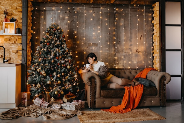 Mujer sentada en el sofá con una taza de café o té junto a un árbol de Navidad en casa